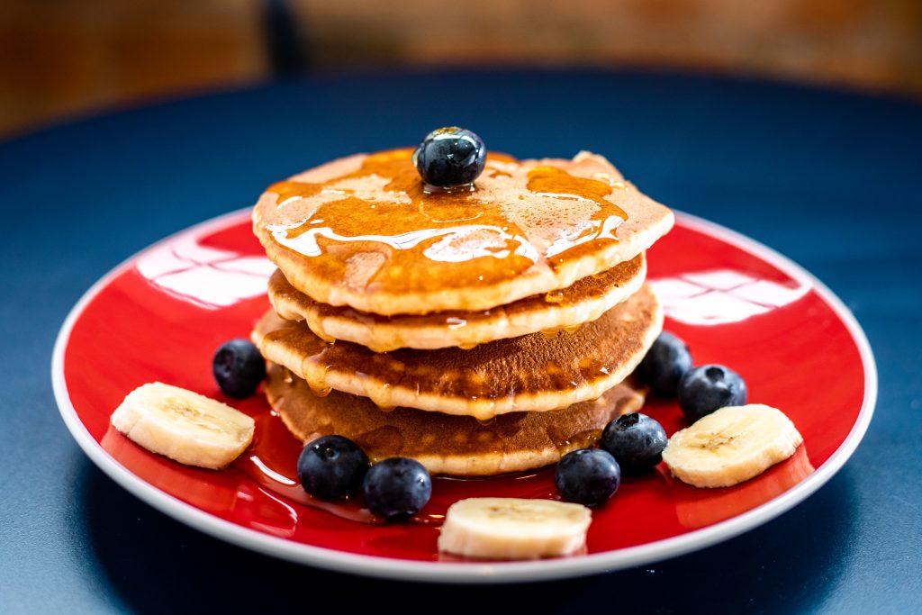 Red plate with four pancakes stacked on top of each other with syrup drizzled on top and blueberries and banana slices